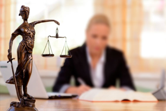 Imagem com o símbolo da justiça. A deusa segurando uma espada e a balança. Ao fundo uma advogada loira escrevendo