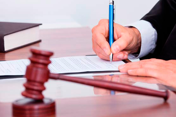 Plano detalhe enquadrando mãos de juiz sobre a mesa assinando um documento, sob a mesa também estão um martelo e um livro