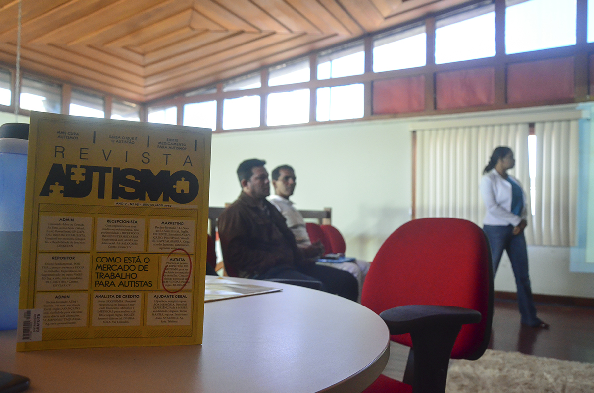 foto de um folder sobre Autismo focado e ao fundo pessoas sentadas e uma mulher de pé