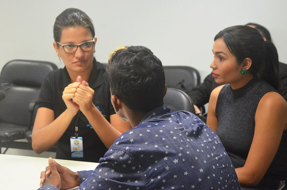 Foto de uma intérprete de libras e gesticulando para um rapaz sentado na sua frente, uma servidora senta ao lado dela