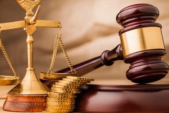 Imagem de uma balança pequena e dourada ao lado de um martelo de juiz (malhete) com seu apoio e entre eles uma pilha de moedas douradas