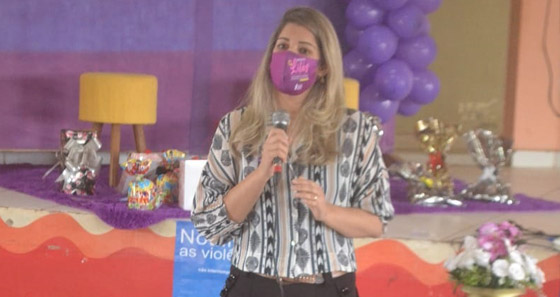 Fotografia da juíza de Direito Louise Kristina, falando ao microfone. Atrás dela tem um palco decorado na cor lilás com bexigas, tapete e presente esparramado por cima do palco.