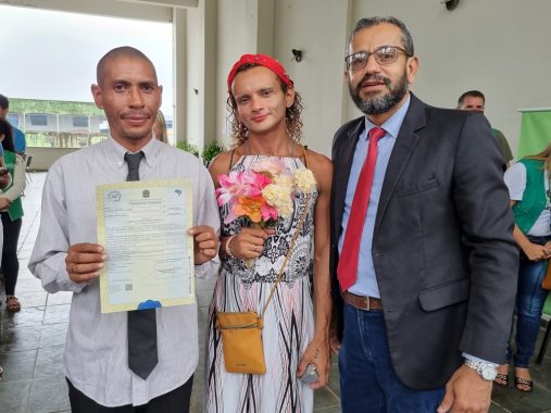 Imagem do juiz Edinaldo ao lado de Jeames e Larissa, segurando a certidão de casamento