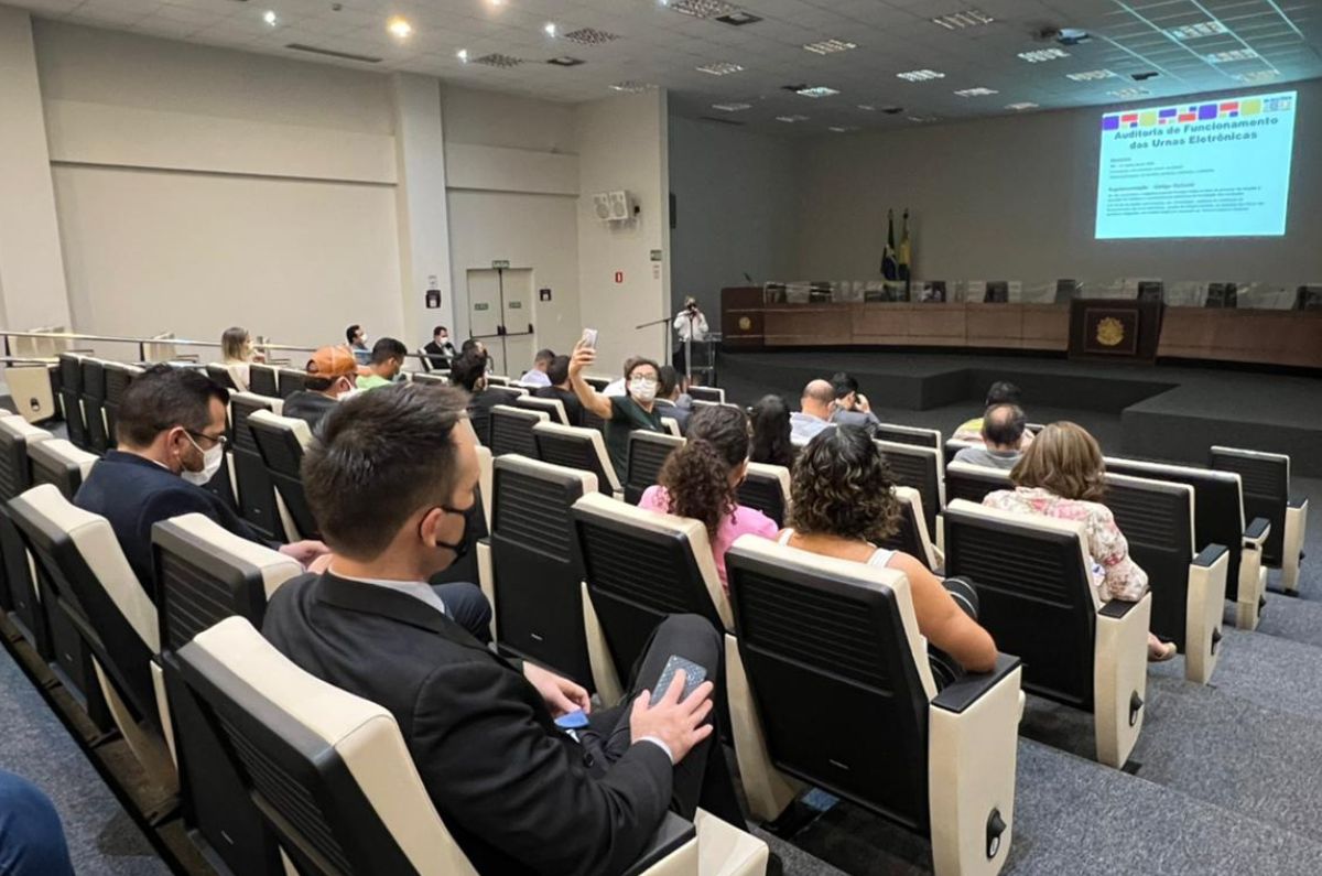 Imagem da plateia sentada olhando para os slides projetado na parede. Ao fundo está uma servidora do TRE falando no microfone