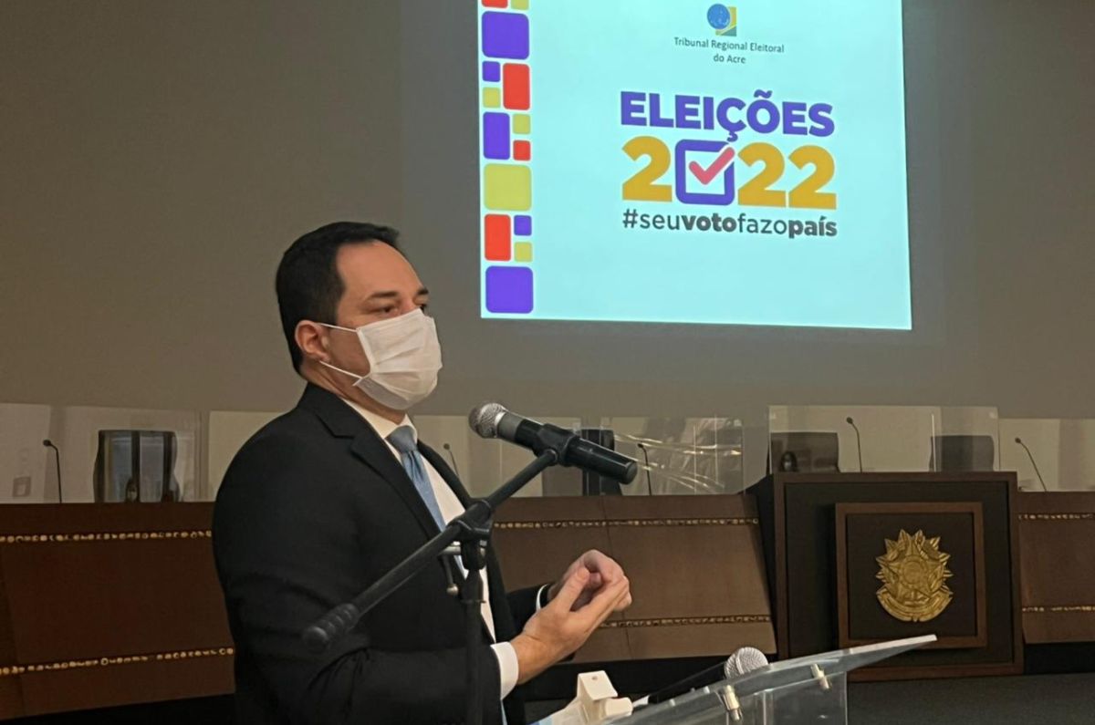 Foto do juiz eleitoral falando no púlpito e atrás aparece um slide projetado na parede com a logomarca do TRE e o texto "Eleições 2022. #seuvotofazopaís
