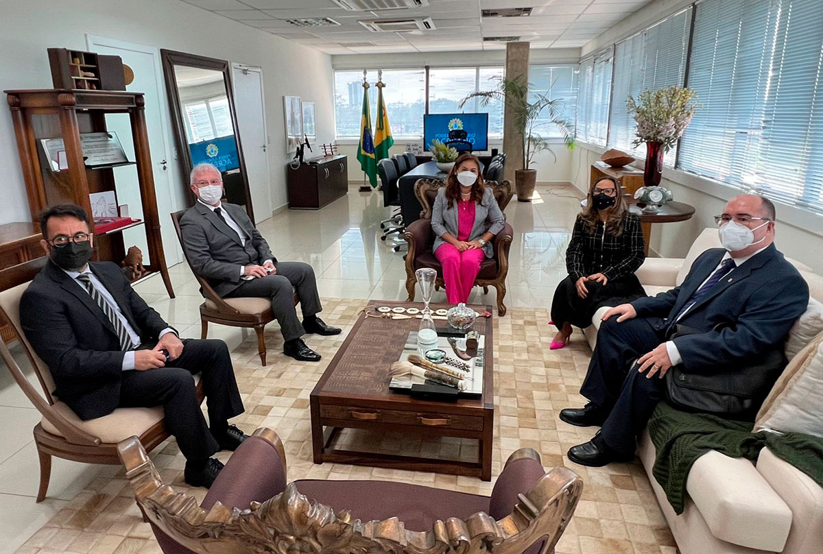 Foto de cinco pessoas sentadas em poltronas e sofás elegantes. Todos olham na direção da câmera. Ao fundo mesas, armários e um espelho grande e retangular