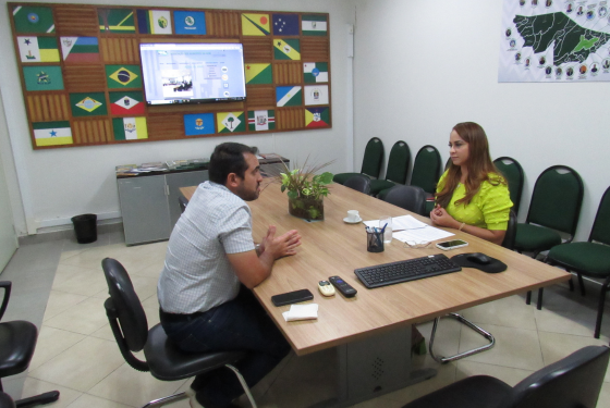 Foto de Isnailda Gondim, servidora da Comsiv, em uma reunião com o coordenador da Amac, Marcus Frederico. Ela está de perfil, usa uma blusa verde, tem cabelos castanhos-alaranjados e está sentada. À sua frente, separados por uma mesa, é possível ver o coordenador da Amac. Ele está de perfil, veste calça jeans e blusa xadrez, seus cabelos são pretos.
