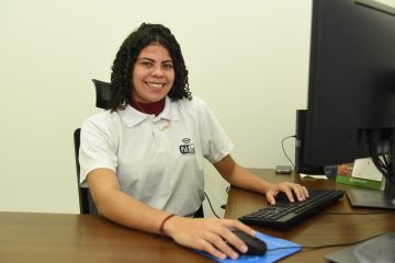 Foto da estagiária do PidJus de Porto Acre, Mariana Malfortes. Ela aparenta ter cerca de 18 anos, é negra, tem cabelos cacheados e usa o uniforme do PidJus.