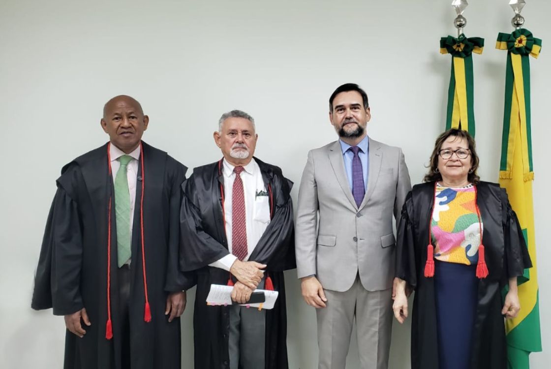 A imagem mostra quatro pessoas posando para uma foto em frente a uma parede branca, com duas bandeiras do Brasil ao fundo, simbolizando um ambiente formal e institucional. Três das pessoas estão vestindo togas pretas com detalhes em vermelho, indicando que são magistrados ou membros do Poder Judiciário. A quarta pessoa, o segundo a partir da direita, veste um terno cinza com camisa azul clara e gravata lilás, sugerindo que também ocupa um cargo de destaque, possivelmente um representante jurídico ou autoridade convidada. A composição da imagem transmite solenidade e profissionalismo, característica de cerimônias ou eventos judiciais, como posses, homenagens ou sessões formais em tribunais.