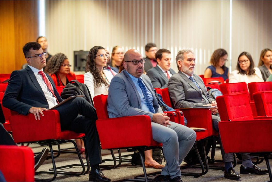 A imagem mostra um auditório com diversas pessoas sentadas em poltronas vermelhas, assistindo atentamente a uma apresentação ou palestra. O ambiente é formal, provavelmente relacionado a um evento institucional, corporativo ou governamental. A maioria das pessoas está vestida com trajes sociais, como ternos e blazers. Em primeiro plano, à esquerda, um homem de terno escuro com gravata vermelha segura um caderno ou bloco de anotações. Ao seu lado, outro homem, de terno azul claro e gravata azul de bolinhas, está descalço, o que chama atenção por contrastar com o contexto formal da situação. Próximo a ele, outro homem, de barba grisalha e terno cinza, segura o que parece ser um tablet ou bloco. Ao fundo, outros participantes — homens e mulheres — também acompanham o evento, alguns com expressões sérias, outros com semblantes mais neutros. Cortinas verticais fechadas e iluminação artificial compõem o ambiente interno. A disposição e o comportamento do público indicam que se trata de um momento de escuta ou discussão séria.