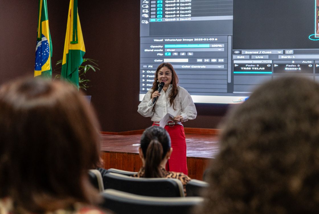 A imagem mostra uma mulher falando em público em um auditório. Ela está em pé sobre um palco, segurando um microfone e algumas folhas de papel, aparentando estar fazendo uma apresentação ou discurso. Detalhes da cena: A mulher veste uma blusa branca e uma saia ou calça vermelha. Ela sorri enquanto fala. Atrás dela, há uma grande tela de projeção com uma interface de software visível, sugerindo que a apresentação envolve o uso de recursos visuais ou tecnológicos. No canto esquerdo da imagem, há duas bandeiras: a do Brasil e a do estado do Acre. Na plateia, vemos algumas pessoas assistindo à apresentação, com foco em uma mulher de cabelos presos no centro. O ambiente é formal e bem iluminado, indicando um evento institucional ou governamental. A imagem transmite um momento de comunicação institucional ou educacional, com foco em troca de informações ou treinamento.