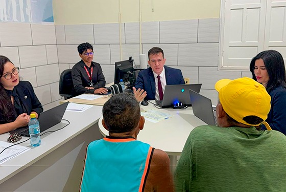 A imagem mostra uma reunião ou atendimento formal em um ambiente de escritório. Seis pessoas estão presentes na cena: Três homens e três mulheres participam do encontro. Duas pessoas, que parecem ser cidadãos buscando atendimento ou consultoria, estão sentadas de costas para a câmera — um usando um boné amarelo e camiseta verde, e o outro com camiseta azul e laranja. Do outro lado da mesa, estão quatro profissionais: duas mulheres e dois homens. Todos parecem estar envolvidos em uma atividade de atendimento jurídico, social ou administrativo. O homem ao centro, de terno e gravata, parece estar liderando a conversa. Ele fala com os dois homens sentados à sua frente e faz gestos com as mãos. Há laptops, documentos e uma garrafa de água sobre a mesa, sugerindo um ambiente de trabalho organizado. Ao fundo, vê-se um ambiente limpo e profissional, com paredes revestidas de azulejo e um quadro ou banner parcialmente visível. A imagem transmite a ideia de um serviço público, mutirão de atendimento ou ação social promovida por alguma instituição governamental ou organização.