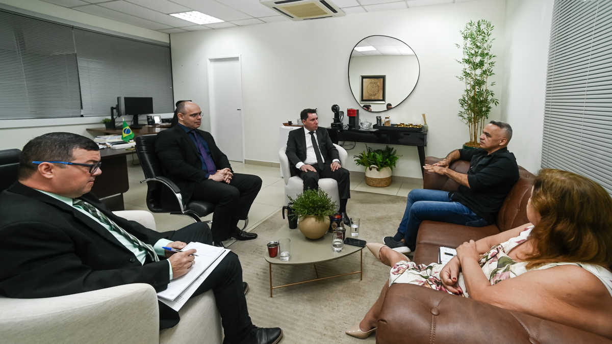 A imagem mostra quatro homens sentados em uma sala de estar ou recepção corporativa, três dos homens usam trajes formais como terno pretos e sapatos social, já o outro homem usa calça jeans e blusa social manga longa preto e tênis preto, já a mulher usa vestido florido com salto alto cor bege e esta com as mãos cruzadas ambos sentados. O ambiente tem um ar elegante e acolhedor, com móveis de madeira esculpida e estofados sofisticados. O homem à esquerda está sentado de forma descontraída em um sofá marrom, com as pernas cruzadas e um sorriso largo no rosto. Ele esta fazendo gesto articulando como se estivesse falando algo.