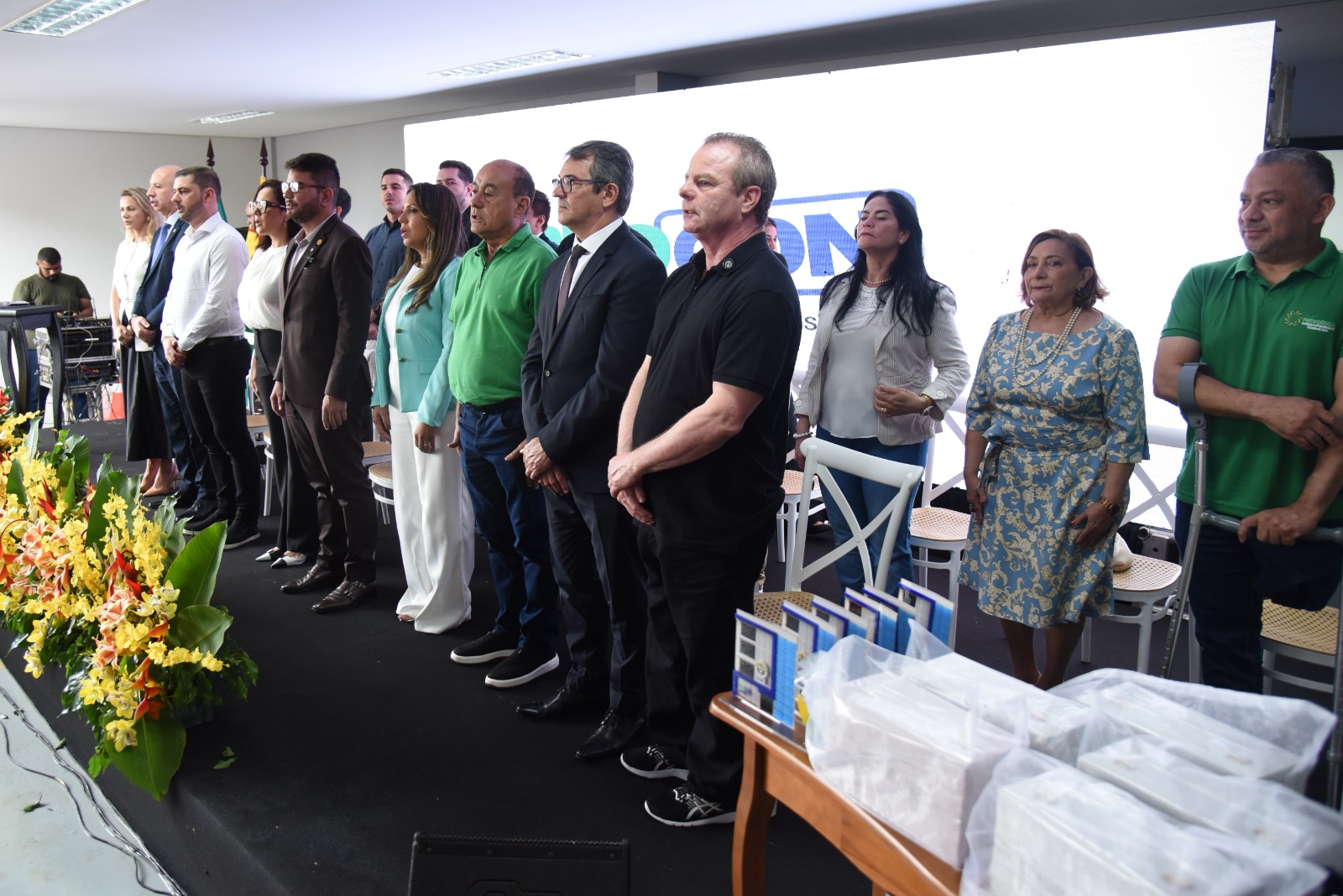 A imagem mostra um grupo de pessoas em um ambiente formal, possivelmente durante uma cerimônia ou evento institucional. Em destaque, estão 11 pessoas entre homens e mulheres posicionadas em pé, ao centro da imagem, sorrindo para a foto. Eles estão elegantemente vestidas — duas usam ternos escuro, um homem veste blusa azul escuro manga de punho e calça bege e tênis preto, já o outro homem veste blusa maga de punho preto e calça preto e tênis a combinar ela esta a esquerda da foto, já o outro vesti blusa ver de manga gola polo e chapéu cor branco, calça jeans azul escuro sapato preto, uma veste um elegante vestido azul com detalhes dourados, e outras três usam roupas brancas. Uma delas segura um pequeno bolsa. Ao fundo, algumas pessoas estão sentadas, entre elas dois homens que também aparentam participar do evento. Um deles usa terno e gravata azul clara, enquanto outro, à direita da imagem, veste uma camisa verde e um chapéu branco, transmitindo uma imagem mais informal. O ambiente é iluminado e moderno, com fundo claro e telão branco, sugerindo um auditório ou sala de cerimônia. A atmosfera geral da imagem é de celebração, reconhecimento ou homenagem, dada a presença do buquê e os sorrisos calorosos dos participantes.