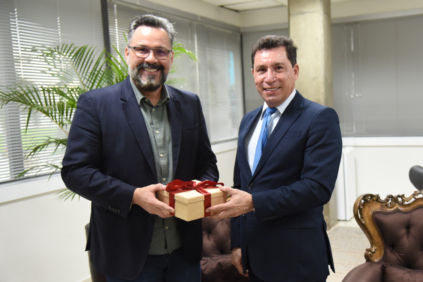 A imagem mostra dois homens em um ambiente corporativo, provavelmente um escritório. Ambos estão vestidos formalmente, usando ternos. O homem à esquerda veste um blazer escuro com camisa verde, enquanto o homem à direita está de terno azul-marinho com camisa listrada e gravata azul. Eles estão sorrindo e segurando juntos uma pequena caixa de presente, que está embrulhada com uma fita vermelha. O gesto sugere uma entrega simbólica de presente ou homenagem, talvez ao final de uma reunião ou evento comemorativo. Ao fundo, é possível ver uma poltrona clássica estofada, uma planta decorativa e janelas com persianas, contribuindo para o ambiente profissional e acolhedor. A atmosfera geral transmite cordialidade, respeito e um clima positivo.