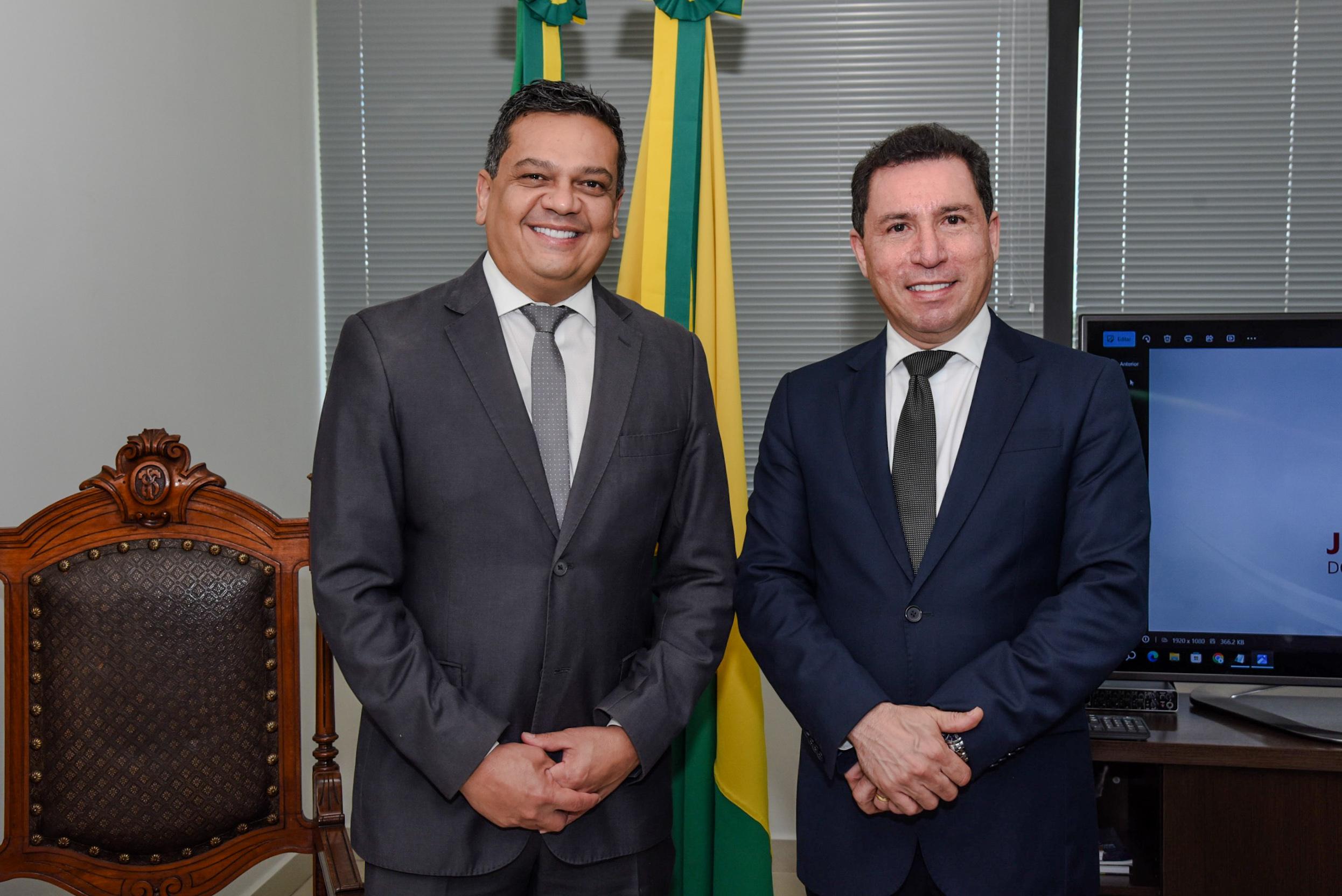 A imagem mostra dois homens em pé, lado a lado, sorrindo para a câmera em um ambiente formal, provavelmente um gabinete institucional ou escritório de autoridade pública. Ambos estão vestidos com ternos escuros, camisa branca e gravata cinza. O homem à esquerda veste um terno cinza escuro, enquanto o da direita usa um terno azul marinho. Atrás deles há duas bandeiras do Brasil posicionadas lado a lado, o que reforça o caráter oficial da ocasião. À esquerda da imagem, aparece parcialmente uma cadeira de madeira entalhada com estofado escuro e detalhes dourados, de estilo mais clássico. À direita, um monitor de computador está ligado e exibe parte de uma apresentação ou documento com o nome “J...”, possivelmente relacionado ao setor jurídico ou institucional, dado o formato da fonte e o layout da tela. A postura dos dois homens, o sorriso e o cenário indicam que se trata de um registro formal e positivo, possivelmente após uma reunião, assinatura de acordo, posse ou visita institucional.