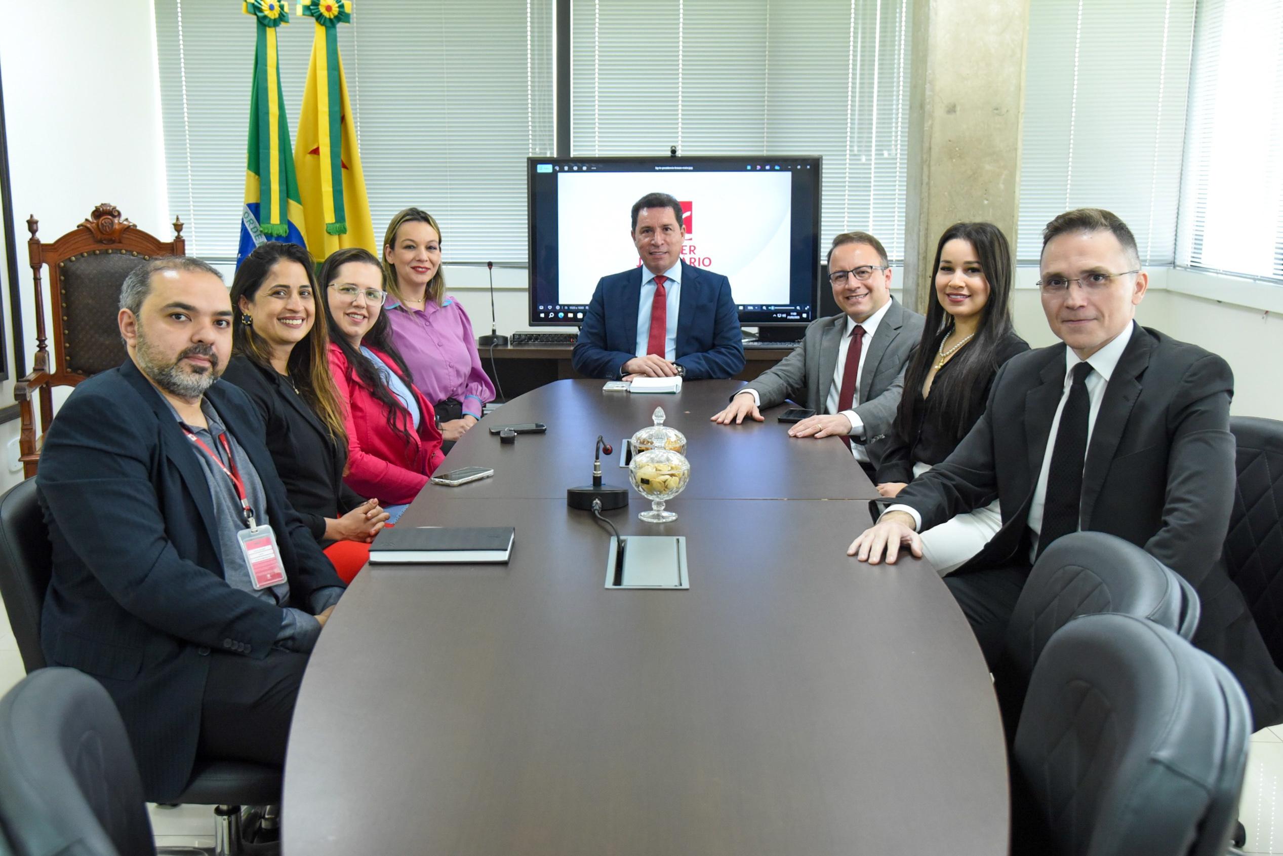 A imagem mostra um grupo de nove pessoas sentadas ao redor de uma mesa de reuniões em um ambiente corporativo ou institucional. Todas estão bem-vestidas, com trajes formais, indicando um encontro profissional ou oficial. Ao fundo, há duas bandeiras — a do Brasil e a do estado do Acre — posicionadas próximas a uma tela ligada, que exibe um logotipo com os dizeres "PODER JUDICIÁRIO". As pessoas estão distribuídas igualmente dos dois lados da mesa, com uma pessoa ao centro da ponta oposta à câmera, sugerindo que possa ser a figura de maior autoridade na reunião. Há objetos sobre a mesa, como celulares, cadernos, um microfone de mesa e dois potes de vidro com balas ou biscoitos.