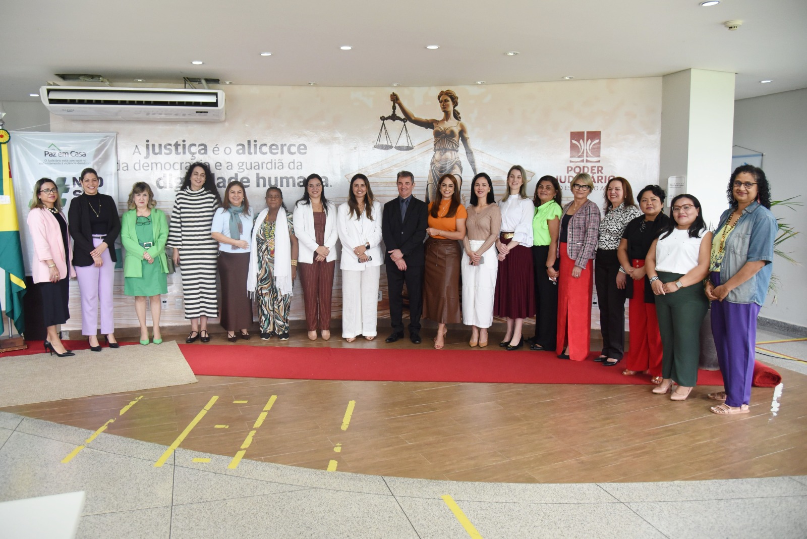 A imagem mostra um grupo de aproximadamente 20 pessoas, majoritariamente mulheres, posando para uma foto em um ambiente institucional. Ao fundo, há um grande painel com a imagem da deusa da justiça (Themis), acompanhada da frase: “A justiça é o alicerce da democracia e a guardiã da dignidade humana”. Também estão visíveis os logotipos do Poder Judiciário do Estado do Acre e da campanha “Paz em Casa”, voltada ao enfrentamento da violência doméstica. As pessoas estão bem-vestidas, com roupas formais e elegantes, sugerindo que participam de um evento oficial, possivelmente ligado à promoção dos direitos das mulheres e à justiça social. O tapete vermelho no chão reforça o caráter solene da ocasião. A composição transmite união, representatividade e compromisso institucional com temas como justiça, dignidade humana e equidade de gênero.