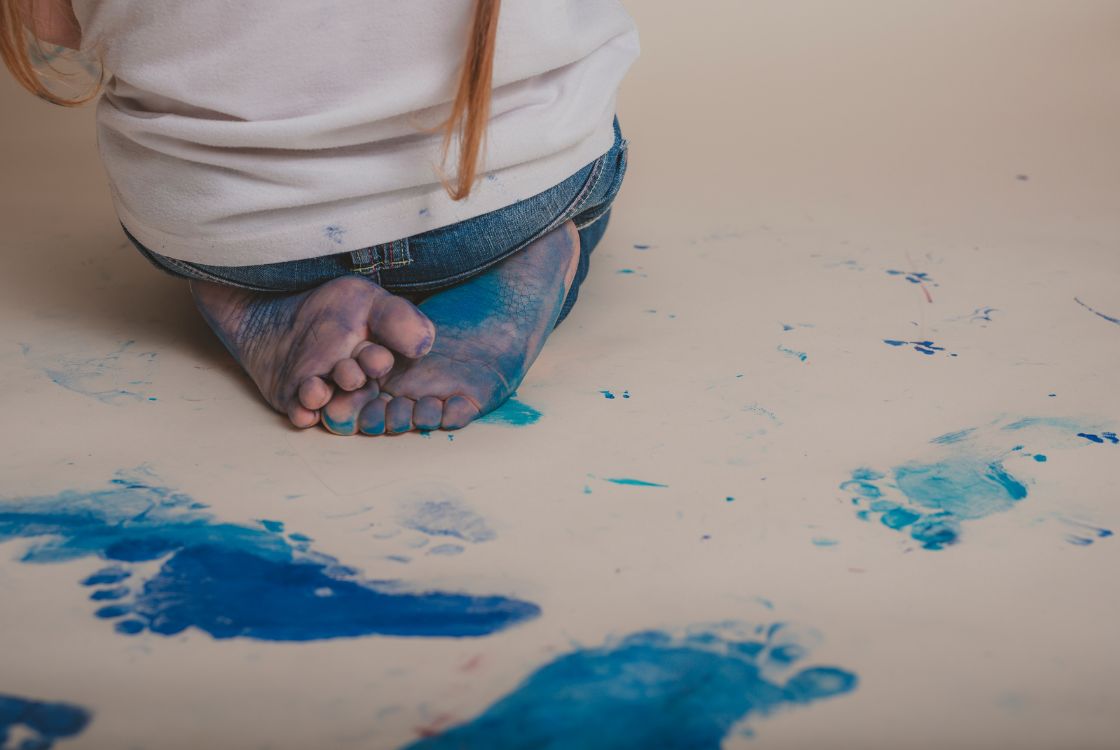 A imagem mostra uma criança ajoelhada no chão com os pés cobertos de tinta azul. Ela está vestindo uma calça jeans e uma camiseta branca. O chão ao redor está coberto de pegadas azuis e manchas de tinta espalhadas. A cena sugere uma atividade artística ou brincadeira com tinta, resultando em um ambiente criativo e descontraído.