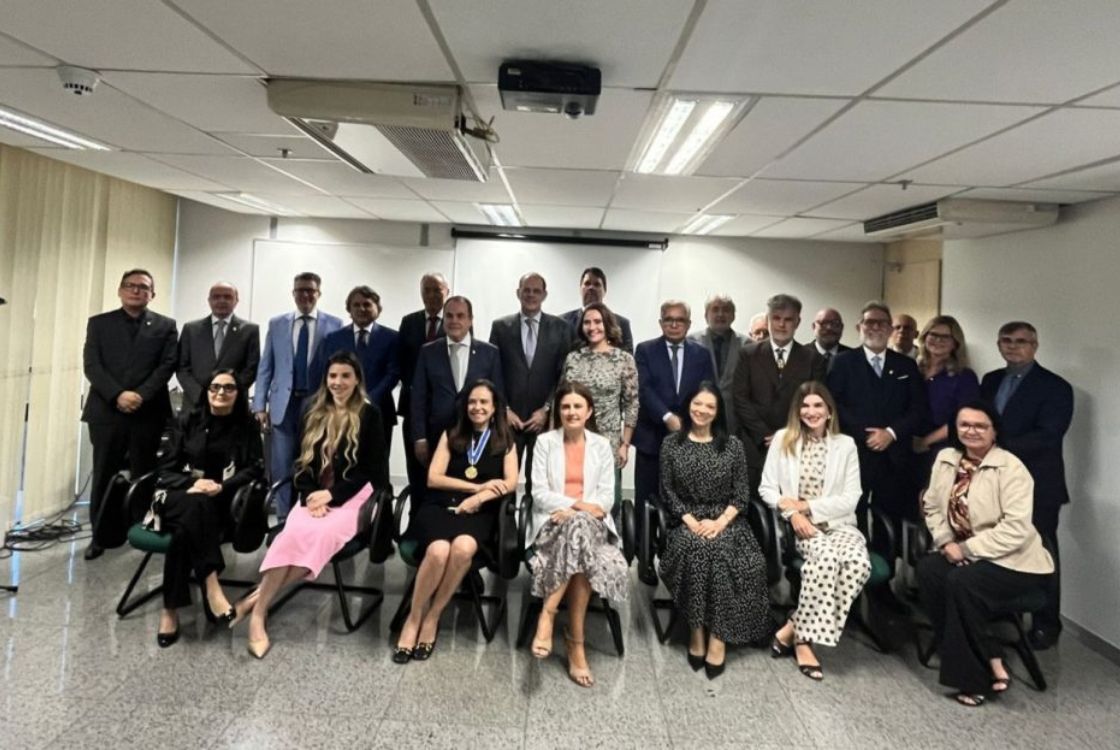 A imagem mostra um grupo de aproximadamente 20 pessoas posando para uma foto em um ambiente corporativo ou institucional. A maioria dos indivíduos veste trajes formais, como ternos e vestidos sociais, sugerindo que se trata de um evento oficial, reunião de trabalho ou cerimônia de premiação. Na primeira fileira, algumas mulheres estão sentadas, incluindo uma delas que usa uma medalha no pescoço, indicando um possível reconhecimento ou homenagem. O fundo contém uma tela de projeção, e há equipamentos audiovisuais no teto, sugerindo que a foto foi tirada em uma sala de conferências ou reunião. A disposição das pessoas e o cenário reforçam a ideia de um evento institucional importante.