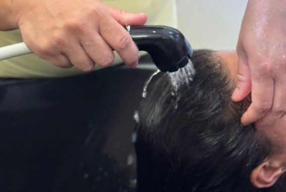 A imagem mostra uma pessoa lavando os cabelos em um lavatório de salão de beleza. Uma mão segura um chuveirinho preto, direcionando a água para os fios molhados da cliente, enquanto a outra mão apoia sua cabeça. O lavatório é preto e possui um formato ergonômico para acomodar o pescoço da pessoa que está recebendo o serviço. A cena transmite um momento de cuidado e higiene capilar, possivelmente parte de um procedimento de lavagem antes de um corte, hidratação ou outro tratamento capilar. A imagem destaca os detalhes das mãos, a água corrente e a textura dos fios molhados.