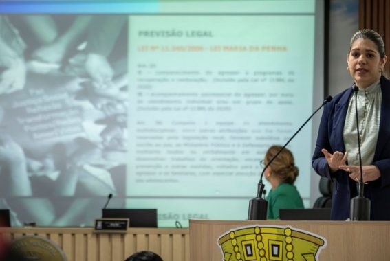 A imagem mostra uma mulher vestindo um blazer azul e uma blusa clara falando em um púlpito com microfone, em um ambiente que parece ser uma câmara municipal ou outro espaço de reuniões oficiais. Ela está gesticulando enquanto discursa, demonstrando expressividade. Ao fundo, há uma apresentação projetada em um telão, com o título "PREVISÃO LEGAL" e a referência à "Lei nº 11.340/2006 – Lei Maria da Penha", sugerindo que o tema abordado seja relacionado à violência doméstica e políticas de proteção às mulheres. Também é possível ver outra mulher, de blusa verde, sentada e voltada para a tela, possivelmente acompanhando a apresentação. O ambiente é formal, com estrutura de madeira clara e monitores sobre a mesa, indicando um espaço utilizado para debates, audiências ou reuniões institucionais.