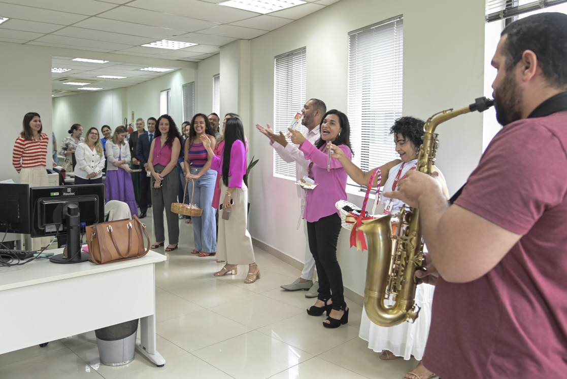 A imagem mostra um momento especial dentro de um ambiente de escritório, onde várias pessoas estão reunidas, aparentemente participando de uma homenagem ou celebração, provavelmente em alusão ao Dia Internacional da Mulher. No primeiro plano, vemos várias mulheres e homens em pé, assistindo atentamente à cena. Algumas pessoas seguram cestas decoradas com laços, provavelmente contendo lembranças ou brindes que estão sendo distribuídos. Um músico com saxofone também está presente, sugerindo que a homenagem incluiu música ao vivo, criando um clima mais afetivo e festivo. O ambiente é moderno, com móveis de escritório, computadores, paredes claras e janelas com persianas, transmitindo uma atmosfera de local de trabalho formal, porém descontraído nesse momento de confraternização. Todos os presentes parecem atentos e envolvidos, demonstrando que foi um momento significativo e de valorização entre colegas.