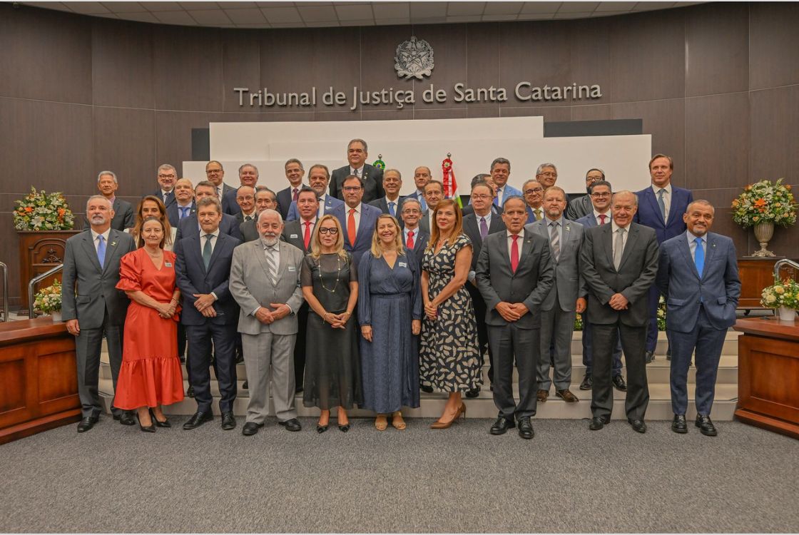 A imagem mostra um grande grupo de pessoas posando para uma foto oficial no Tribunal de Justiça de Santa Catarina. São homens e mulheres vestidos formalmente, com ternos e vestidos, dispostos em várias fileiras. O ambiente tem uma decoração com arranjos de flores ao fundo e o brasão do tribunal acima do nome da instituição.