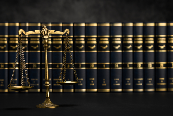 A imagem mostra uma balança dourada da justiça em destaque, com um fundo composto por livros jurídicos encadernados em preto e dourado. A iluminação cria um efeito sofisticado e sério, remetendo ao universo do direito e da advocacia. Esse tipo de composição é frequentemente utilizado para representar conceitos como equidade, julgamento e conhecimento jurídico.