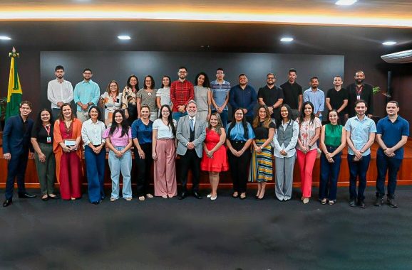 imagem mostra um grande grupo de pessoas posando para uma foto oficial em um ambiente institucional. O grupo é diverso, composto por homens e mulheres vestidos de forma formal e semi-formal, com roupas sociais, vestidos, calças e camisas. Algumas pessoas usam crachás, indicando que podem ser funcionários ou participantes de um evento corporativo ou governamental. O fundo da imagem apresenta um painel escuro, uma bandeira do Brasil posicionada no canto esquerdo e uma iluminação sofisticada no teto, sugerindo que a foto foi tirada em um auditório ou sala de conferências. O grupo está distribuído em dois níveis, com algumas pessoas em pé sobre uma plataforma elevada e outras no chão, organizadas de forma equilibrada para que todos apareçam na foto. A expressão das pessoas é, em geral, positiva, com sorrisos e posturas descontraídas, indicando um momento de celebração ou encerramento de um evento significativo.