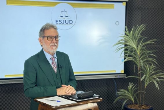 A imagem mostra um homem com cabelos grisalhos e barba, usando óculos e um terno verde com um colete combinando, falando em microfone. Ele está de pé atrás de um pódio de madeira e parece estar fazendo um discurso ou palestra. No fundo, um vaso de planta é visível, indicando que o cenário pode ser formal ou oficial. Há também uma tela exibindo uma imagem do símbolo da esjud. A atmosfera sugere um evento profissional ou cerimonial.