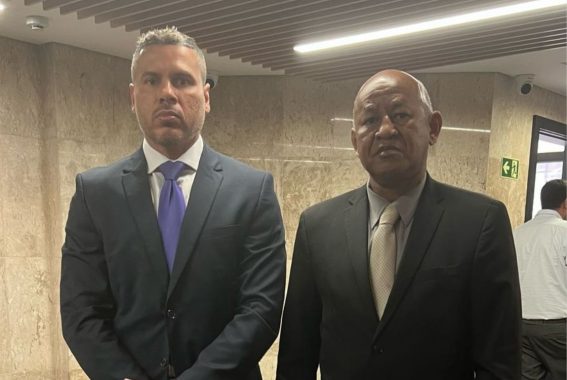 Na imagem, dois homens estão sérios posando para uma foto, um deles tem cor de pele branca, cabelo e barba branca, usando um terno preto, camisa social branca e gravata roxa, o outro homem tem cor de pele preta, careca, usa terno preto, camisa social cinza e gravata bege escuro