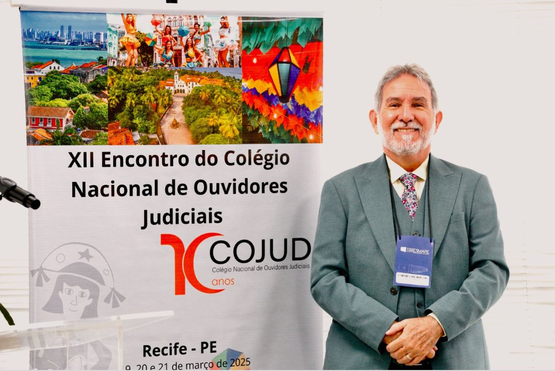 A imagem mostra um homem sorridente, de barba grisalha, vestindo um terno verde, camisa branca e gravata com estampa floral. Ele usa um crachá azul pendurado no pescoço e está posicionado ao lado de um banner do "XII Encontro do Colégio Nacional de Ouvidores Judiciais" (COJUD), que celebra seus 10 anos. O banner contém imagens de Recife, incluindo pontos turísticos, manifestações culturais e elementos do carnaval pernambucano, como o frevo e bandeirolas coloridas. No canto inferior, há a ilustração de um personagem típico do Nordeste brasileiro, possivelmente representando um cangaceiro estilizado. O evento está programado para acontecer nos dias 20 e 21 de março de 2025, em Recife-PE. Ao lado do homem, há um púlpito de acrílico com um microfone. O ambiente tem iluminação clara, com fundo branco e persianas.