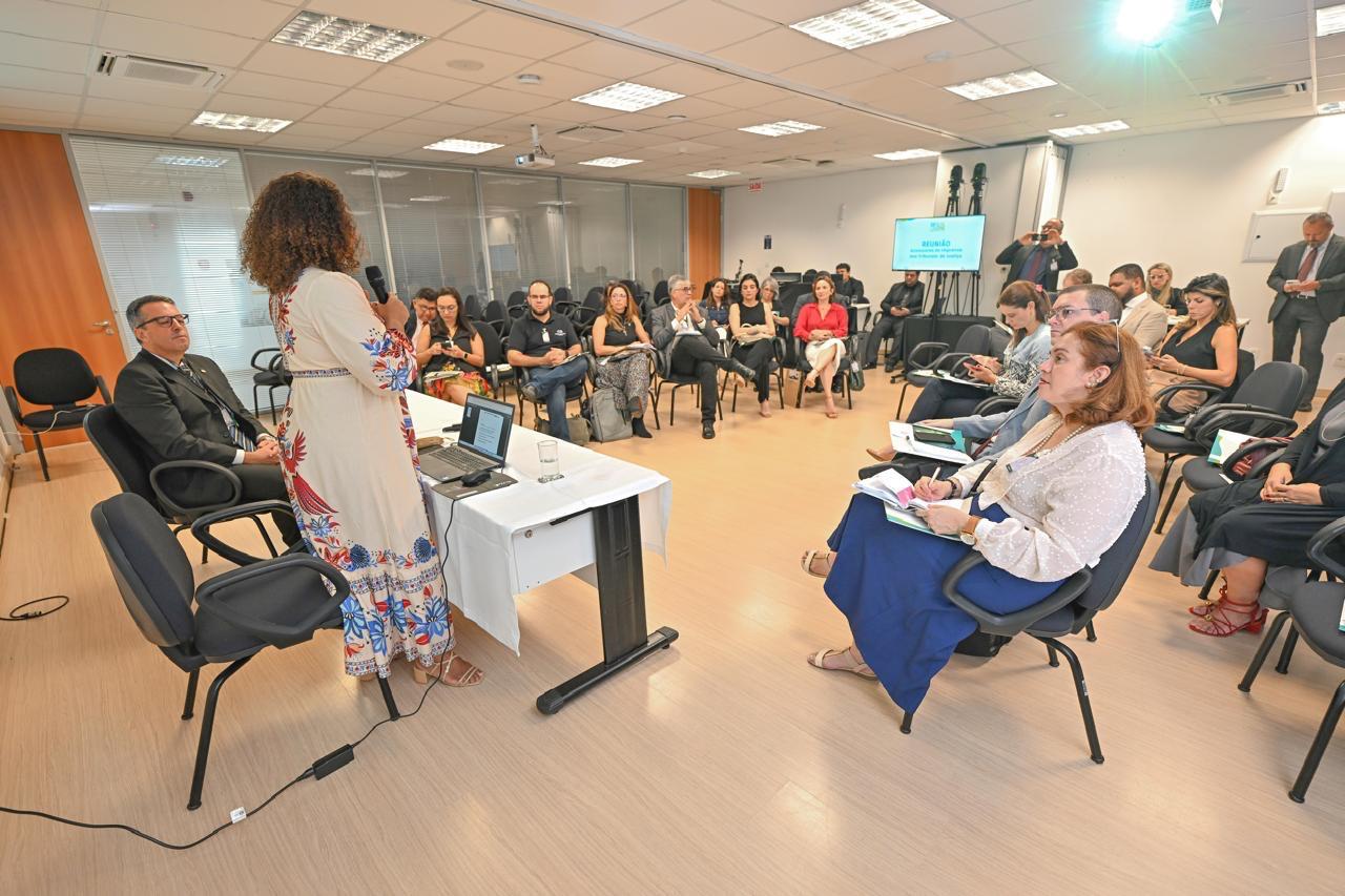 Uma mulher está fazendo uma apresentação em uma sala com várias pessoas sentadas em cadeiras, ouvindo atentamente. A composição da cena mostra uma sala de reuniões ou conferência, com cadeiras dispostas em círculo em torno de uma mesa onde uma palestrante está localizada no centro da imagem. As pessoas sentadas estão concentradas na palestrante, sugerindo uma sessão de apresentação ou palestra. O enquadramento é amplo, mostrando o ambiente geral da sala e a interação entre a palestrante e os ouvintes. A palestrante está voltada para a audiência e em frente a um local para apresentação. As posições das pessoas sugerem um momento de aprendizado ou de escuta atenta. A palestrante é uma mulher adulta com cabelo castanho, em uma postura atenta e segura. Ela está usando um vestido estampado, provavelmente leve e confortável para o ambiente. A audiência é constituída por pessoas de diferentes idades, com diferentes tipos de vestimentas. As expressões faciais da audiência sugerem atenção e interesse na palestra. O meio técnico da imagem é uma fotografia convencional, tirado de um ângulo que mostra a maioria dos participantes. A qualidade da imagem é boa, sem distorções significativas, e os detalhes são bem visualizados. O estilo é neutro, focado na representação precisa da cena e das pessoas que estão nela. A sala é iluminada por luz natural ou artificial, com iluminação uniforme que não apresenta sombras bruscas ou contrastes fortes. As paredes e o piso são de tons claros e neutros, criando um ambiente profissional e formal. O ambiente parece ser um espaço público para reuniões ou palestras. A atmosfera é de concentração e profissionalismo, com o enfoque principal na apresentação e na audiência.