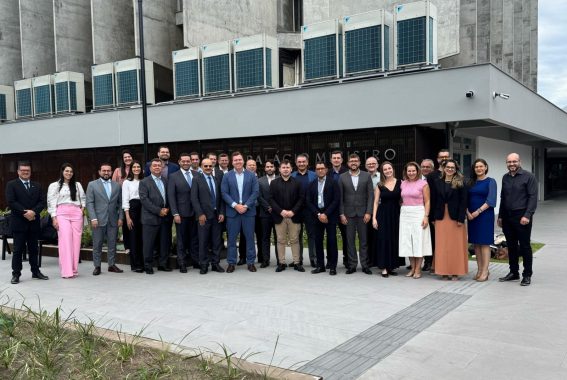 A imagem mostra um grupo de aproximadamente 25 pessoas posando para uma foto em frente a um edifício moderno com a inscrição “PALACIO MESTRO” na fachada. O grupo é composto por homens e mulheres vestidos formalmente, com muitos usando ternos e gravatas, enquanto algumas mulheres optam por vestidos ou saias. A atmosfera parece institucional, possivelmente uma reunião de trabalho, evento governamental ou conferência. O fundo do prédio tem estruturas de ar-condicionado visíveis, e o ambiente externo conta com áreas ajardinadas e um piso moderno. A expressão das pessoas na foto é geralmente positiva, sugerindo um momento de confraternização ou registro de um evento importante.