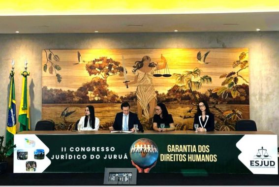 A imagem mostra uma mesa com quatro pessoas sentadas em um evento jurídico. Ao fundo, há um grande painel de madeira com uma ilustração da deusa da justiça segurando a balança e a espada, cercada por elementos da natureza, como árvores e pássaros. Na frente da mesa, um banner exibe o título do evento: "II Congresso Jurídico do Juruá" e o tema "Garantia dos Direitos Humanos", além do logotipo da ESJUD (Escola do Poder Judiciário do Acre). À esquerda, estão bandeiras do Brasil e do estado do Acre.