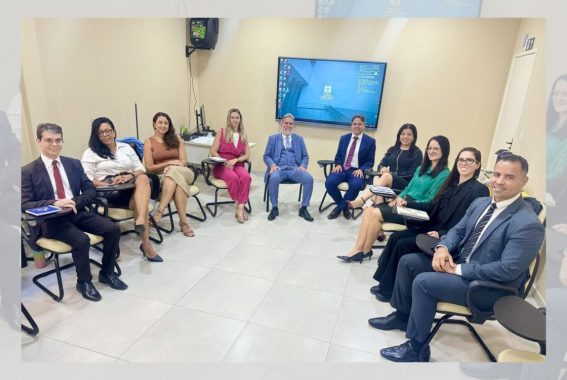 A imagem mostra um grupo de dez pessoas sentadas em círculo em uma sala de reuniões. A sala possui um ambiente profissional, com paredes claras e uma tela grande ao fundo exibindo a área de trabalho de um computador. As pessoas estão vestidas de forma formal ou semiformal, com ternos, blusas sociais, vestidos e saias. Algumas seguram cadernos ou anotações, sugerindo uma reunião ou discussão profissional. O grupo é diverso, composto por homens e mulheres de diferentes idades e estilos. Todos estão sorrindo e parecem engajados na atividade.