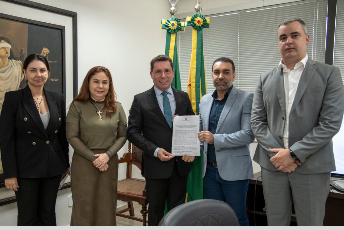 A imagem mostra um grupo de cinco pessoas posando para uma foto em um ambiente formal, provavelmente um gabinete ou escritório governamental. No centro, um homem de terno preto e gravata azul segura um documento oficial, enquanto um outro homem, de terno azul claro, também aponta para o papel. Atrás deles, há duas bandeiras verde e amarela com adornos no topo, sugerindo um contexto institucional ou governamental. As outras três pessoas na foto são duas mulheres à esquerda e um homem à direita. Uma das mulheres veste um blazer preto e a outra, uma blusa verde com acessórios dourados. O homem à direita veste um terno cinza e uma camisa branca. Todos têm expressões sérias e parecem estar participando de um momento de assinatura ou entrega de um documento importante. Ao fundo, há um quadro com a imagem de uma figura feminina vendada segurando uma balança, um símbolo clássico da Justiça. O ambiente tem janelas com persianas fechadas, uma mesa e cadeiras ao redor, reforçando a formalidade do encontro.