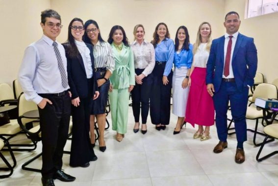 A imagem mostra um grupo de nove pessoas posando para uma foto em um ambiente interno que se assemelha a uma sala de reuniões ou auditório. O grupo é composto por homens e mulheres vestindo trajes formais, sugerindo um evento profissional, reunião ou treinamento. As cadeiras ao fundo são do tipo universitário, com pranchetas acopladas, e há um livro grande sobre uma delas, possivelmente um material de estudo ou referência jurídica. As expressões dos participantes são amigáveis e receptivas, com todos olhando para a câmera. O grupo apresenta diversidade de estilos, com algumas pessoas usando ternos e outras optando por roupas formais com cores variadas. O ambiente é bem iluminado, com paredes claras e um design simples.