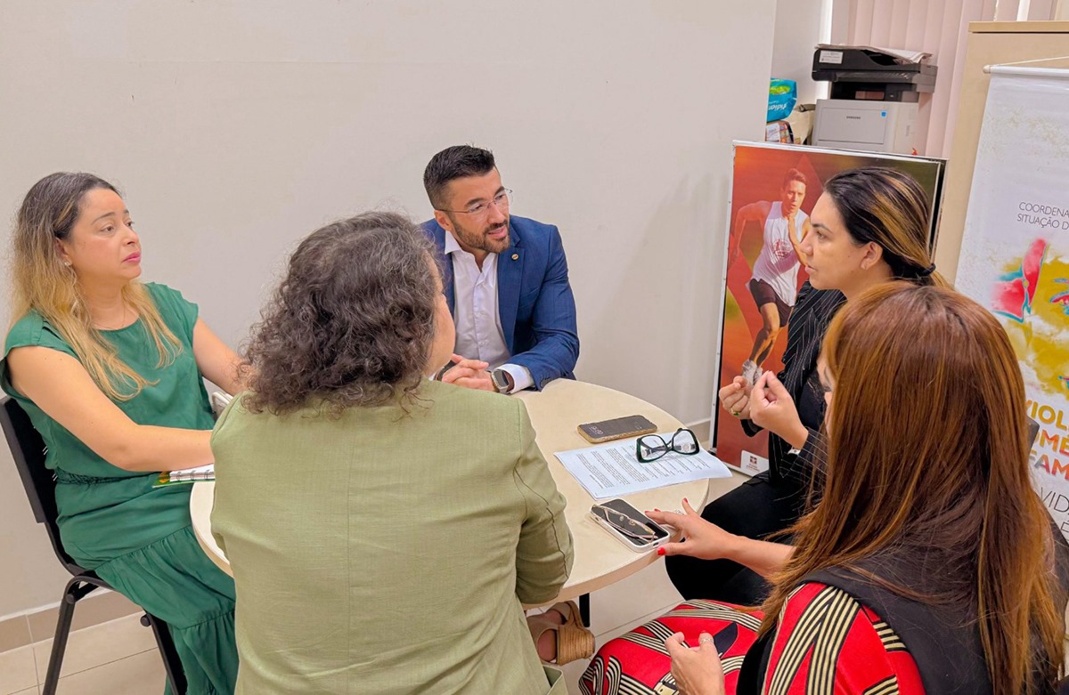 A imagem mostra um grupo de cinco pessoas, quatro mulheres e um homem, reunidas ao redor de uma mesa redonda em um ambiente interno, possivelmente uma sala de reunião ou escritório institucional. A conversa parece séria e focada, sugerindo uma discussão ou planejamento de ações, talvez relacionadas a políticas públicas ou projetos sociais. Alguns elementos reforçam essa ideia: Sobre a mesa há documentos, celulares e um par de óculos, indicando que estão em uma reunião formal. Ao fundo, há um banner parcialmente visível com a frase "Violência contra a mulher e familiar", associado à Coordenadoria da Mulher em Situação de Violência Doméstica e Familiar, o que sugere que o tema da reunião pode estar relacionado ao enfrentamento da violência doméstica. Também é visível um cartaz com a imagem de um corredor, o que pode indicar a presença de materiais de campanhas institucionais diversas no local. As pessoas estão bem vestidas e atentas, reforçando o caráter profissional e engajado da conversa.