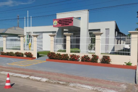 A imagem mostra a fachada de um edifício institucional, identificado como o Fórum Waldemir Jardim Alves Ferreira, pertencente ao Poder Judiciário do Acre, na Comarca de Rodrigues Alves. A construção apresenta um design moderno e bem conservado, com muros pintados de branco e detalhes em bege. Há um portão gradeado, canteiros floridos e uma área de calçada acessível, incluindo uma vaga demarcada para pessoas com deficiência. As bandeiras estão hasteadas a meio mastro, o que pode indicar um período de luto oficial.