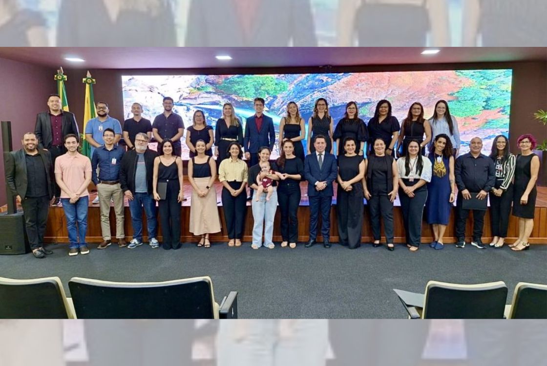 A imagem mostra um grupo de pessoas posando para uma foto em um auditório. O grupo é diversificado, com homens e mulheres vestidos de maneira formal e semiformal. Alguns estão de preto, outros com roupas mais claras. No centro, há um homem de terno e uma mulher segurando um bebê. Ao fundo, há uma tela grande exibindo uma imagem de paisagem natural e duas bandeiras do Brasil. A foto parece ter sido tirada em um evento oficial ou cerimônia institucional.
