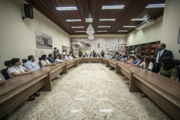 A imagem mostra um grupo de pessoas sentadas em uma sala de reuniões institucional. Ao fundo, há um grande painel com imagens históricas e a inscrição "Poder Judiciário do Estado do Acre", indicando que o evento está relacionado ao sistema judiciário dessa região. A disposição das mesas em formato de "U" e a presença de crachás, copos de água e documentos sugerem que a foto foi tirada ao final de uma reunião ou cerimônia formal. O grupo é diverso, composto por homens e mulheres de diferentes idades, vestidos com trajes formais e semi-formais, demonstrando um ambiente profissional. A presença de bandeiras institucionais e a iluminação sofisticada, com lustres no teto, reforçam o caráter solene do evento. O número de participantes e o posicionamento deles indicam que se trata de um momento de celebração, reconhecimento ou encerramento de um encontro de importância para o Poder Judiciário do Acre.