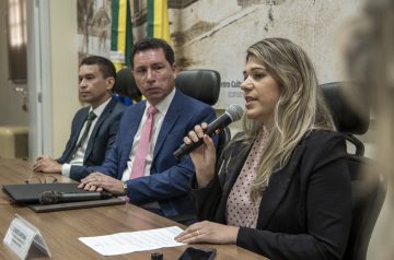 A imagem mostra uma reunião formal em uma sala com prateleiras cheias de livros ao fundo. No centro, um homem de óculos, terno azul e gravata rosa segura um microfone enquanto fala com uma expressão séria e gesticula com a mão. Ele está sentado à mesa ao lado de outro homem de terno verde e gravata verde, que aparece em primeiro plano, levemente desfocado. Ao fundo, uma mulher com roupas formais está de pé em um púlpito, parecendo acompanhar a discussão. Outra mulher, vestindo uma roupa estampada, está sentada à mesa e observa a conversa com um leve sorriso. A mesa está organizada com papéis, pastas, copos de água e placas de identificação, indicando que se trata de uma reunião oficial ou um debate institucional. O ambiente sugere um espaço de decisão jurídica, política ou administrativa.