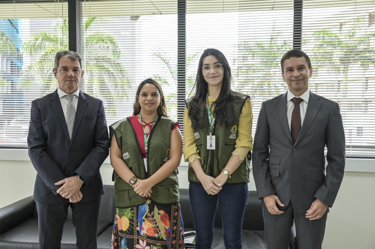 A imagem mostra quatro pessoas posando para uma foto em um ambiente corporativo ou governamental. Duas delas estão vestidas com coletes verdes contendo identificações e logotipos, sugerindo que fazem parte de uma organização pública ou de fiscalização. As outras duas pessoas estão vestindo ternos formais, demonstrando um contexto profissional. O grupo está alinhado e posicionado de forma séria, mas com expressões amigáveis, especialmente o homem à direita, que sorri levemente. No fundo, há janelas grandes com persianas abertas, permitindo a entrada de luz natural e mostrando uma paisagem com palmeiras e prédios. O ambiente parece ser um escritório ou sala de reuniões, coerente com o cenário das imagens anteriores.