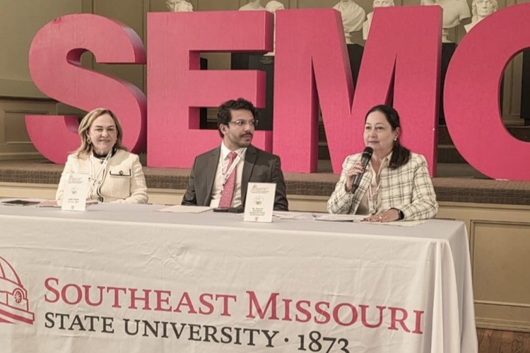 A imagem mostra três pessoas sentadas à mesa em um evento na Southeast Missouri State University, como indicado pela inscrição na toalha branca que cobre a mesa. Atrás delas, há grandes letras vermelhas formando a sigla "SEMO". Os participantes parecem estar em uma conferência ou painel de discussão. A mulher à direita está segurando um microfone e falando, enquanto os outros dois estão ouvindo atentamente. O homem no centro veste um terno e gravata, e as duas mulheres estão com trajes formais, uma delas usando um blazer claro. O ambiente sugere um evento acadêmico ou institucional.