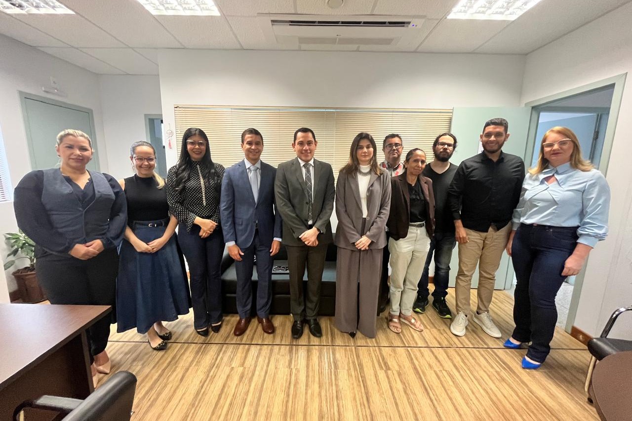 A imagem mostra um grupo de onze pessoas posando para uma foto em um ambiente corporativo ou institucional. Eles estão dispostos em uma única fileira, olhando para a câmera e sorrindo. O ambiente tem paredes claras, móveis modernos e um piso de madeira. Algumas pessoas estão vestidas formalmente, com ternos e roupas sociais, enquanto outras têm um traje mais casual. O fundo da imagem apresenta uma janela com persianas fechadas e uma mesa com cadeiras ao redor. A disposição e o contexto sugerem que o grupo pode estar participando de uma reunião ou evento institucional.