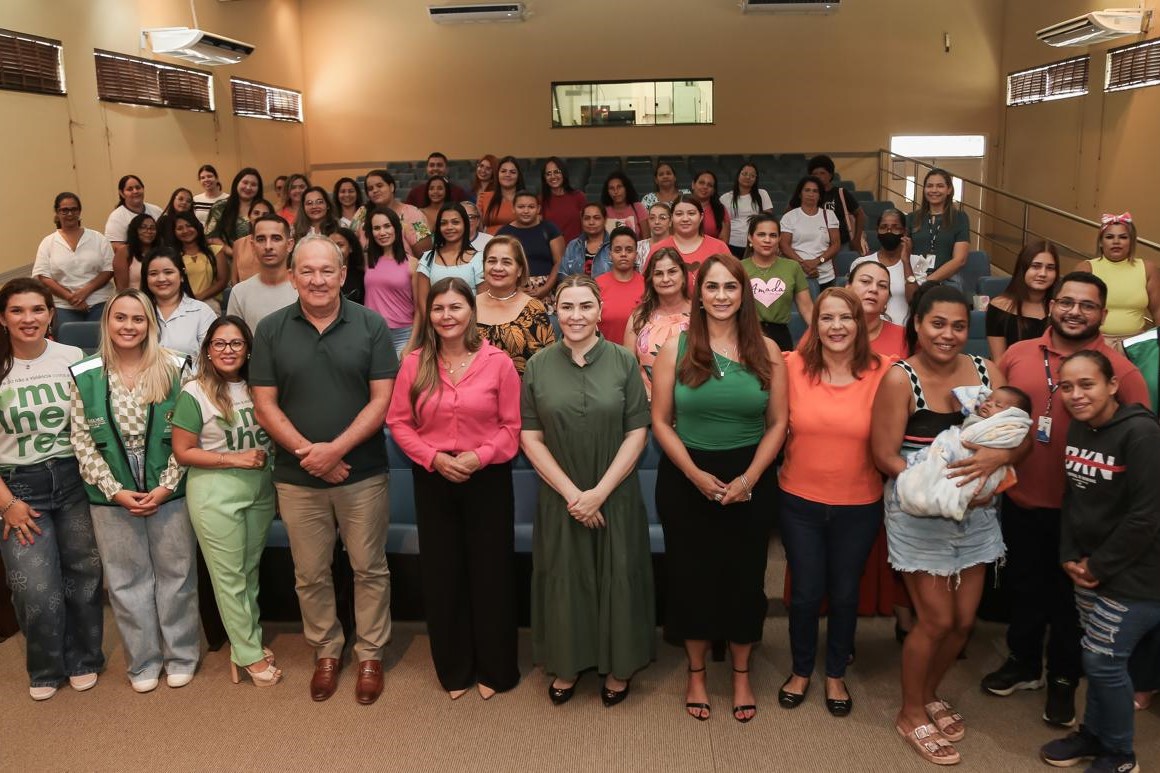 A imagem mostra um grupo grande de pessoas reunidas em um auditório. Na frente, há uma fileira com cerca de 13 pessoas posando para a foto — homens e mulheres, alguns com roupas sociais, outros com roupas mais casuais. Ao fundo, várias outras pessoas estão sentadas ou em pé, completando o ambiente com um clima de encontro ou evento comunitário. Entre os participantes, há mulheres com camisetas temáticas (como "mulheres" e outras estampas), uma mulher segurando um bebê, e pessoas com crachás, indicando possível envolvimento institucional ou organizacional. O ambiente sugere uma reunião, roda de conversa ou ação social voltada ao público feminino, possivelmente relacionada a temas como saúde, assistência social, direitos das mulheres ou políticas públicas. O clima da imagem transmite acolhimento, diversidade e engajamento social.