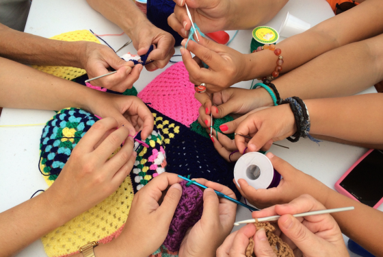 A imagem mostra várias mãos trabalhando juntas em um projeto de crochê. Cada pessoa segura uma agulha de crochê e está tecendo fios de diferentes cores. Há peças coloridas já confeccionadas sobre a mesa, com padrões vibrantes em tons de amarelo, azul, rosa e preto. Alguns acessórios de costura, como um carretel de linha e uma fita métrica, também estão presentes. Os pulsos de algumas pessoas têm pulseiras e adornos, e há uma grande diversidade de tons de pele, sugerindo um ambiente colaborativo e inclusivo. A cena transmite um clima de cooperação e criatividade.