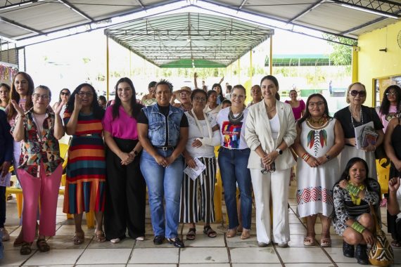 A imagem mostra um grupo diverso de mulheres reunidas em um espaço coberto, possivelmente durante um evento comunitário ou social voltado à promoção de direitos, inclusão ou celebração cultural. O ambiente tem cadeiras amarelas e piso de azulejo, e parece ser uma escola ou centro comunitário. As mulheres estão em pé, posando para a foto com expressões alegres, demonstrando união e empoderamento. Elas estão vestidas de maneira variada, com roupas casuais, sociais e tradicionais — destacando-se uma mulher com vestimenta indígena com colares coloridos e outra com vestido com estampas étnicas, o que reforça a representatividade e diversidade cultural do grupo. O evento aparenta valorizar a inclusão de diferentes etnias e idades, promovendo visibilidade e protagonismo feminino em um contexto de valorização social.